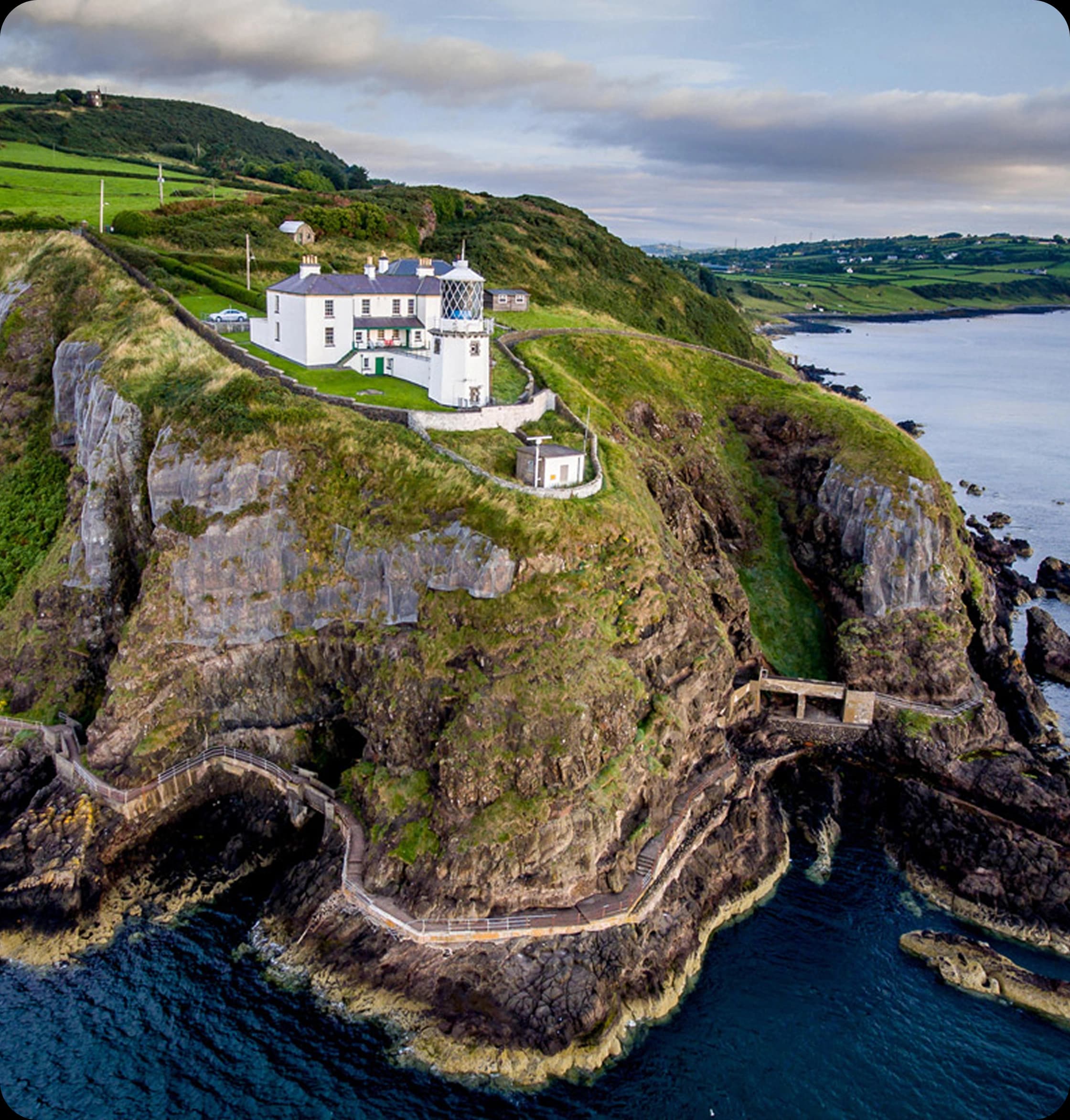 A white house with a lighthouse on a green cliff overlooking the ocean with a winding path